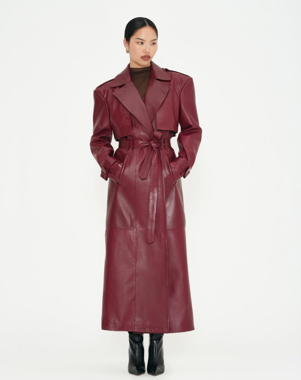 Model wearing a burgundy leather trench coat with a tied belt, black turtleneck, and black knee-high boots against a white background.