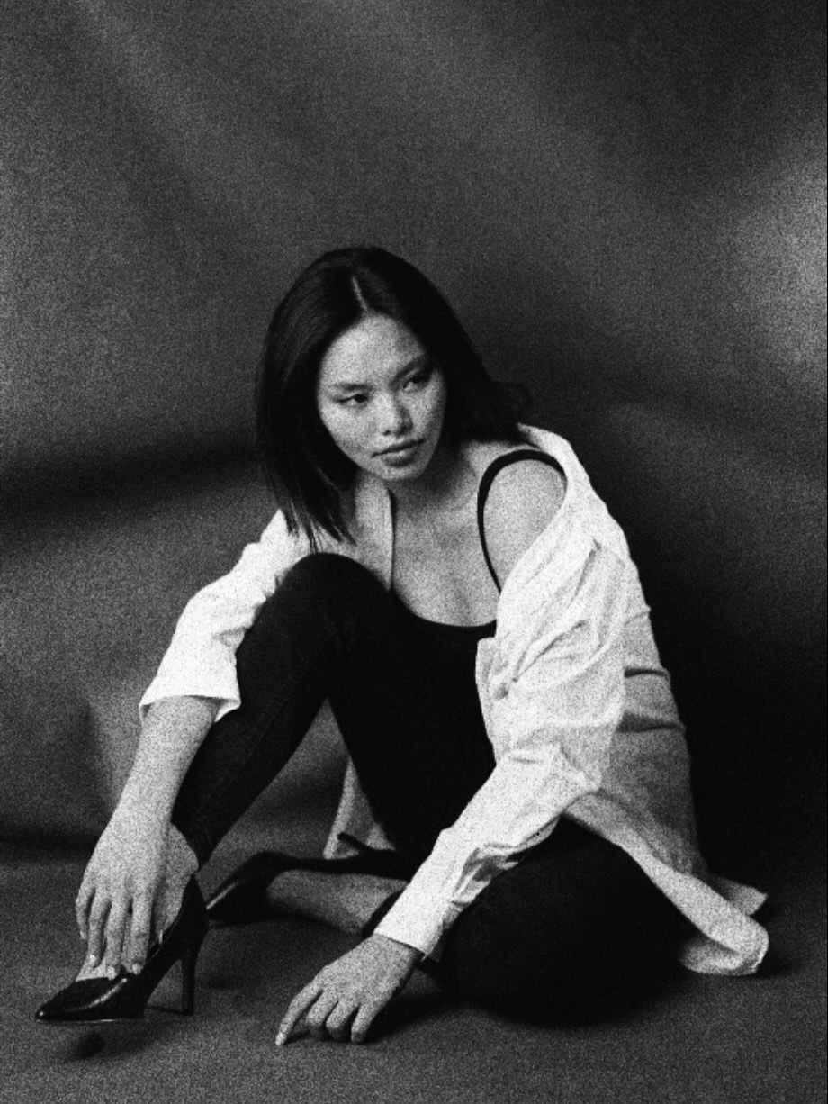 Monochrome photo of a woman seated on the floor, one knee up, wearing a white shirt over a black top and dark jeans, looking to the side.