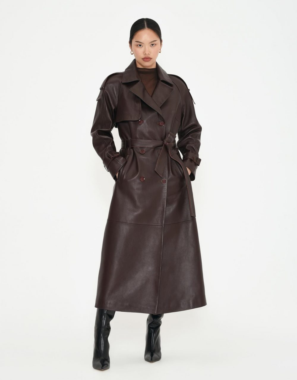Woman modeling a knee-length dark brown leather trench coat with a belt and tall boots against a white backdrop.