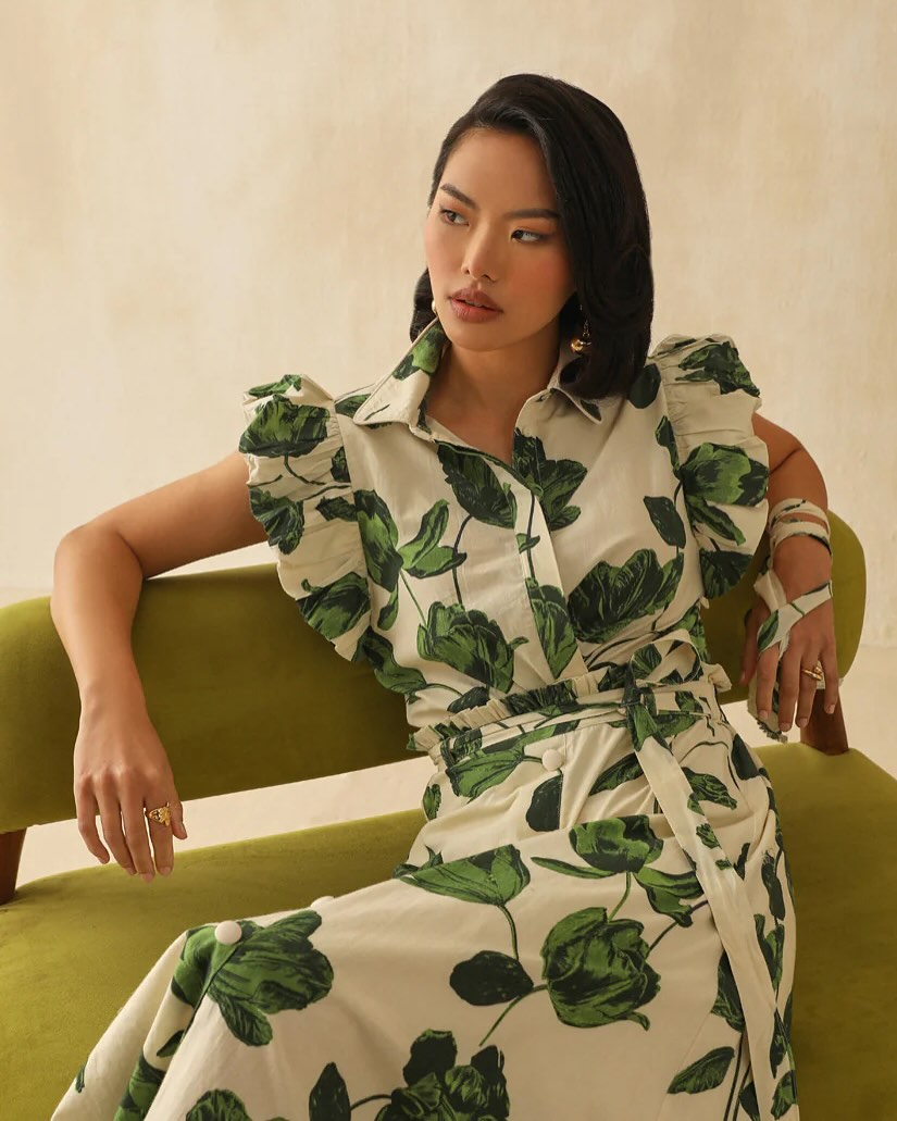 Fashion model seated on a green sofa wearing a white dress with large green leaf print and ruffled sleeves.