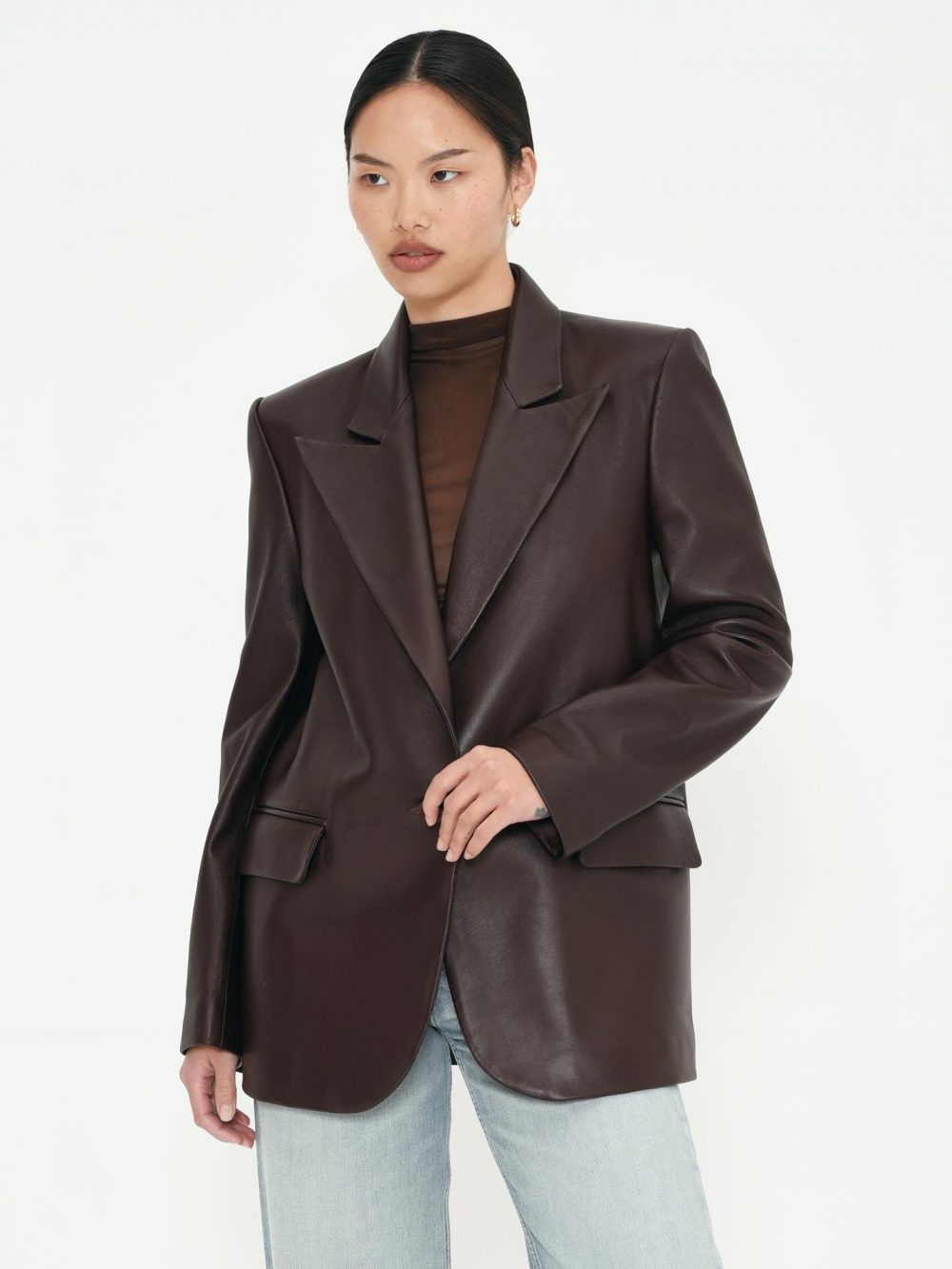 Woman wearing a dark brown leather blazer over a sheer brown top and light denim jeans against a white background.