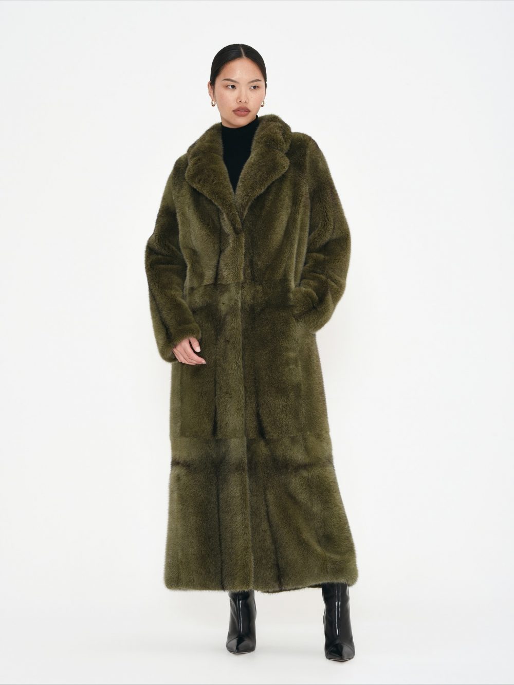 Person standing in a long olive-green fur coat over a black turtleneck with black knee-high boots.