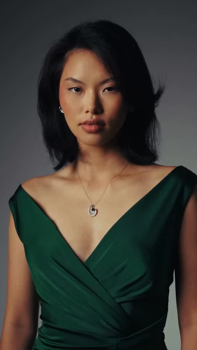 Portrait of a woman in a deep green V-neck dress with a pendant necklace, against a dark studio backdrop, looking slightly to the side.