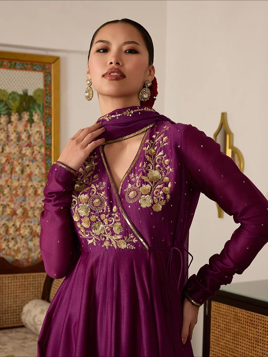 Woman in a purple embroidered dress strikes a confident pose, hand at her neck, wearing large gold earrings.