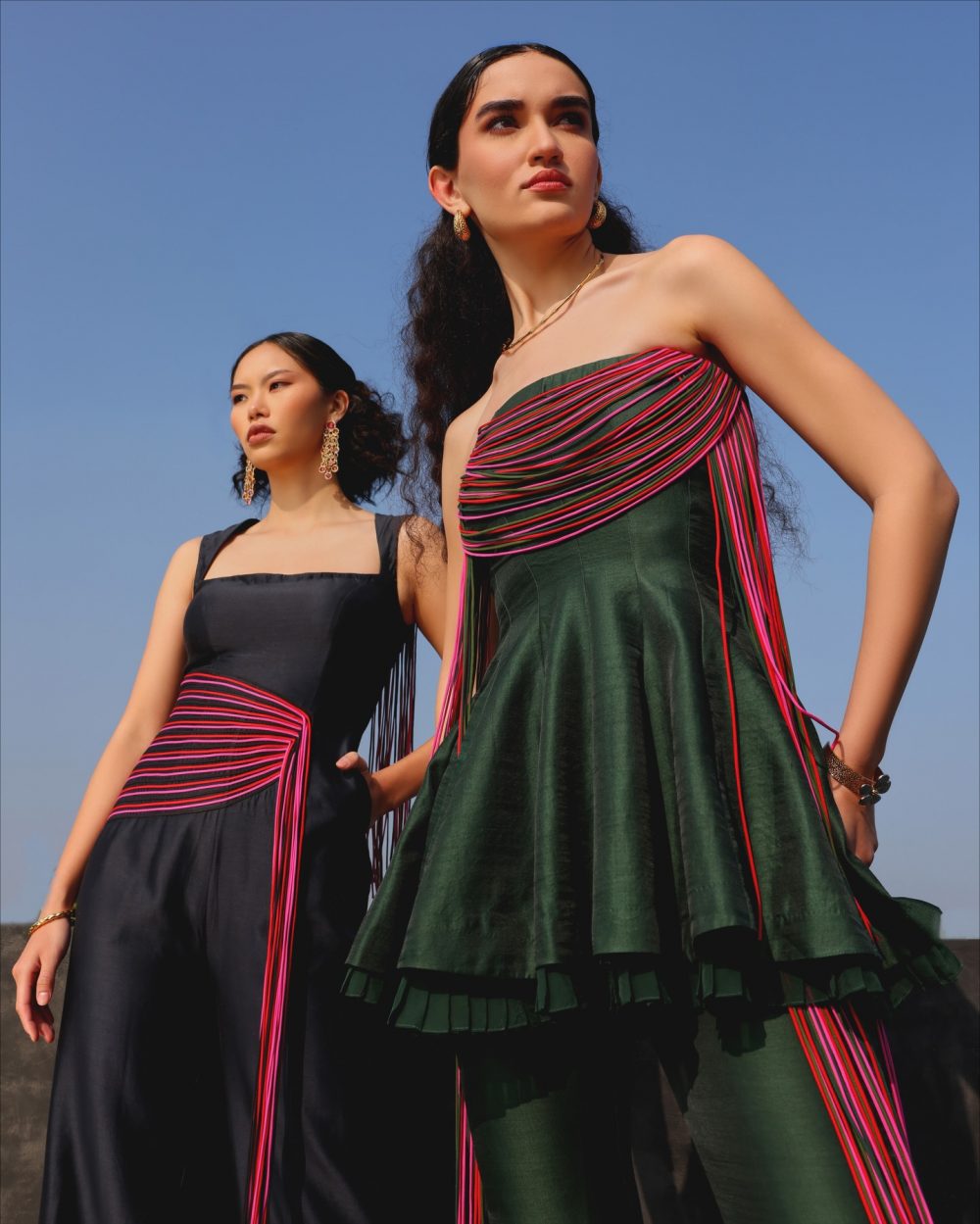 Two models pose in stylish dresses with pink fringe details against a clear blue sky, showcasing bold fashion.