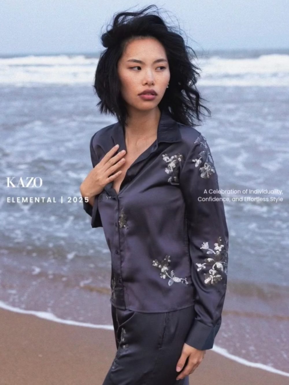 Fashion model on a windy beach wearing a dark silvery blouse with floral embroidery, posing for a KAZO Elemental 2025 campaign.