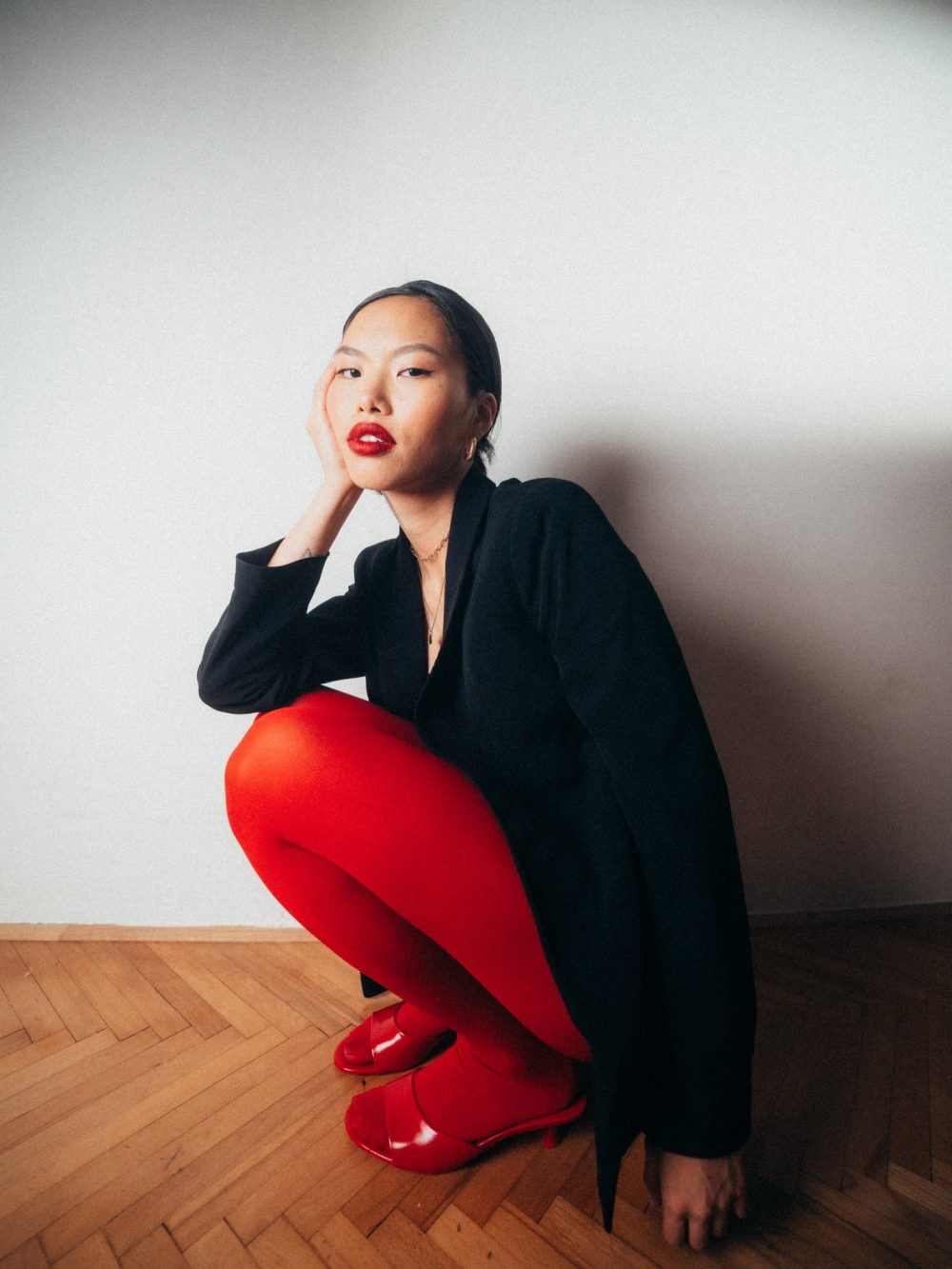 Portrait of a woman wearing a black blazer and bright red pants, squatting by a wall with her hand resting on her cheek, red lipstick visible.