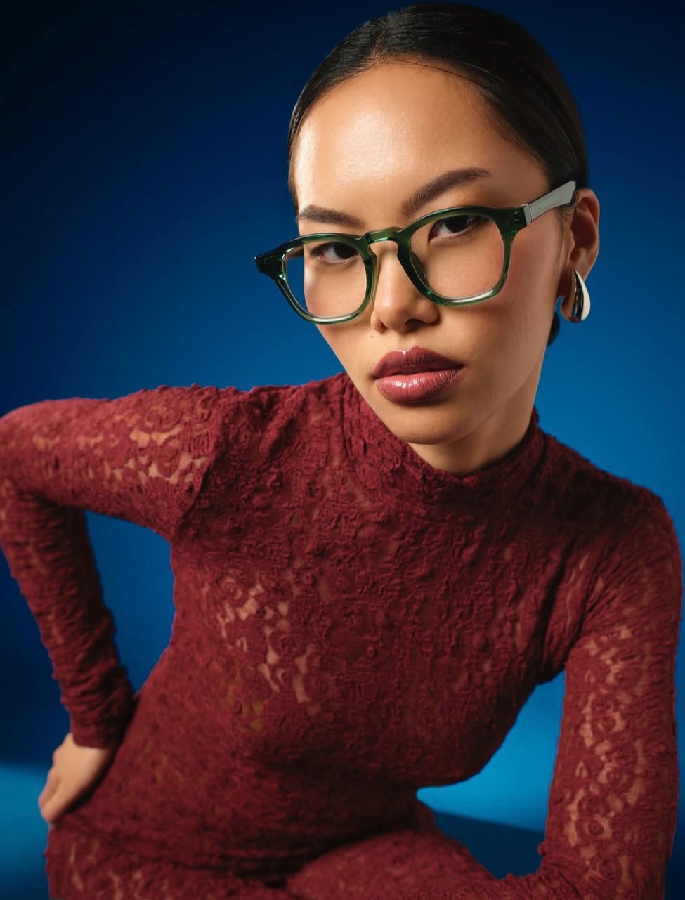 Portrait of a woman with dark hair in a burgundy lace dress, wearing bold green glasses, posed against a blue gradient background.