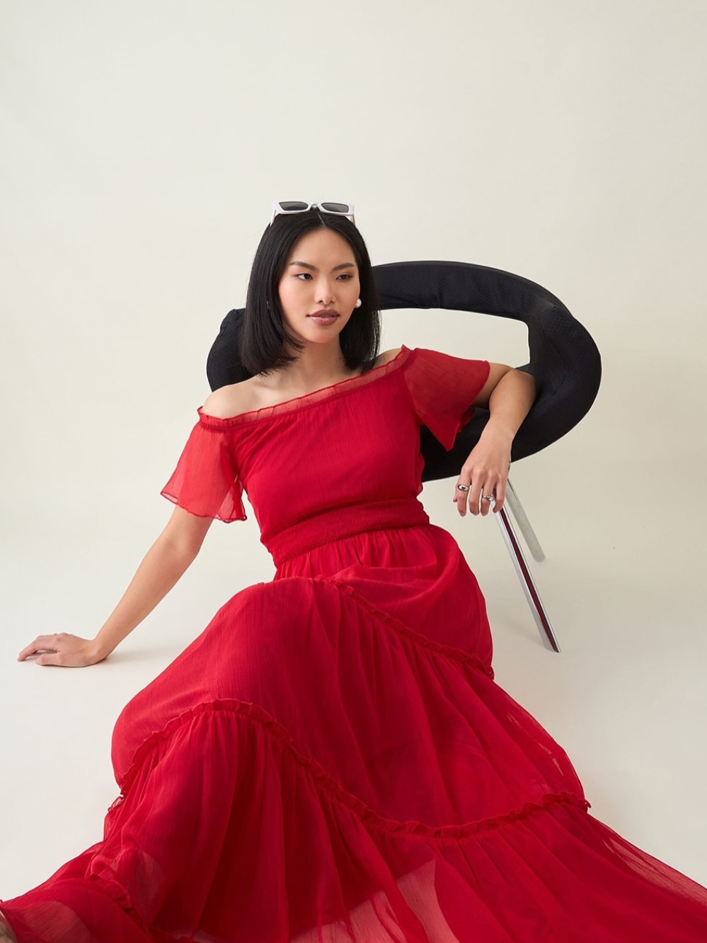 Woman in a bright red off-shoulder dress, seated on a black curved chair against a light background, looking to the side.