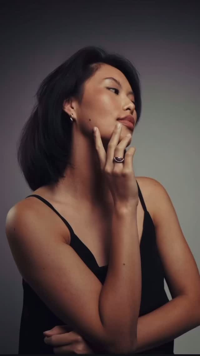 Woman with short black hair wearing a black spaghetti-strap top, touching her face in a thoughtful pose