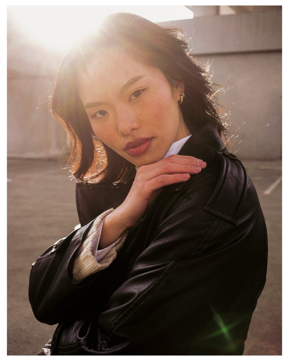Portrait of a woman outdoors with strong sun glare behind her, hands resting near her chin in a black jacket.