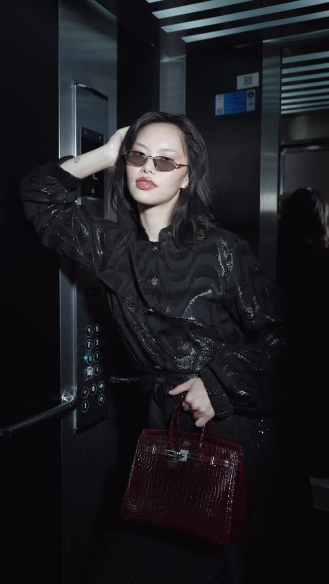 Woman in a black patterned jacket with sunglasses leans against elevator wall, holding a burgundy crocodile‑embossed handbag.