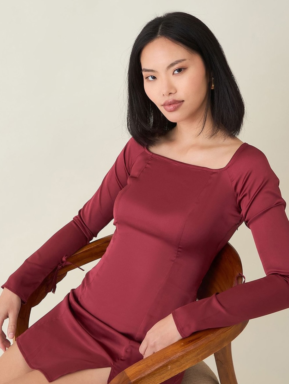Woman in a burgundy long-sleeve dress seated in a wooden chair, looking at the camera against a neutral backdrop.