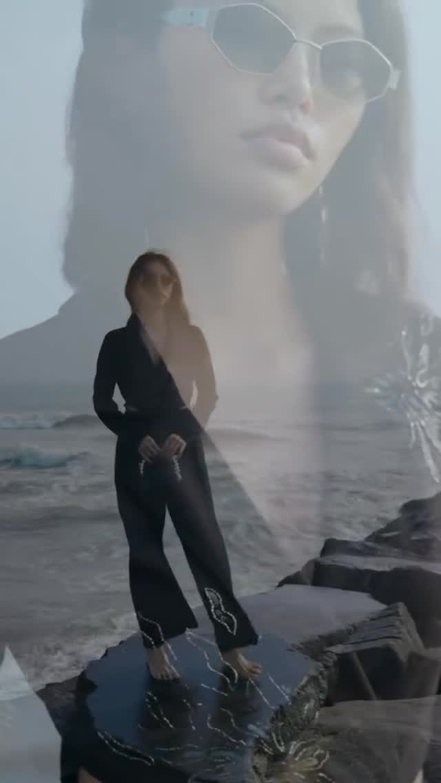 Double-exposed image: a woman in a black jumpsuit standing on rocks by the ocean, overlaid with a large, semi-transparent portrait wearing sunglasses.