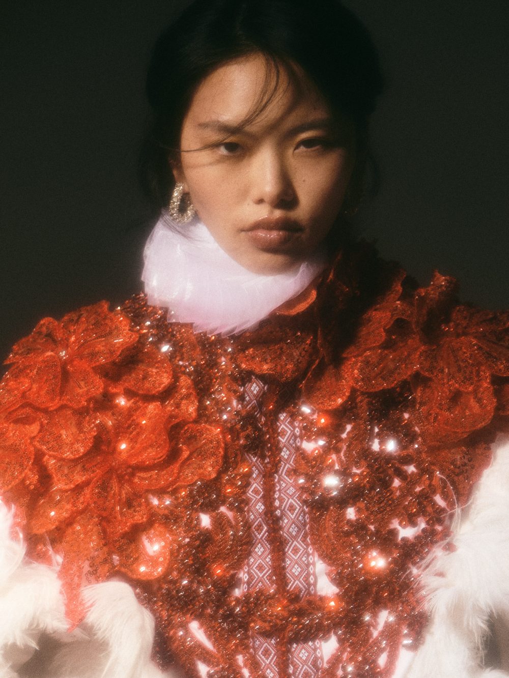 Portrait of a woman with dark hair wearing a red sequined outfit and white ruffled collar, accessorized with large earrings, looking to the side in soft lighting.