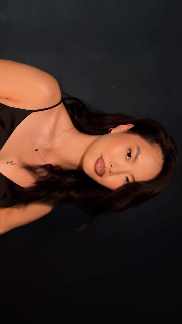 Portrait of a woman with long dark hair, wearing a black top and a gold pendant necklace against a dark background.
