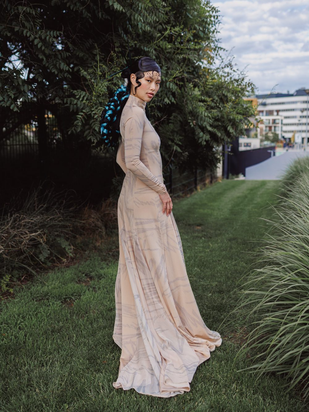 Woman in a long beige patterned dress stands in a grassy garden, looking over her shoulder with blue braided hair.