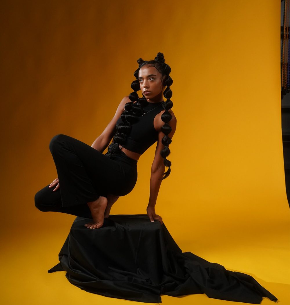 Model in black sleeveless top and pants crouches on a covered pedestal against a bright yellow studio backdrop, with long coiled braids.