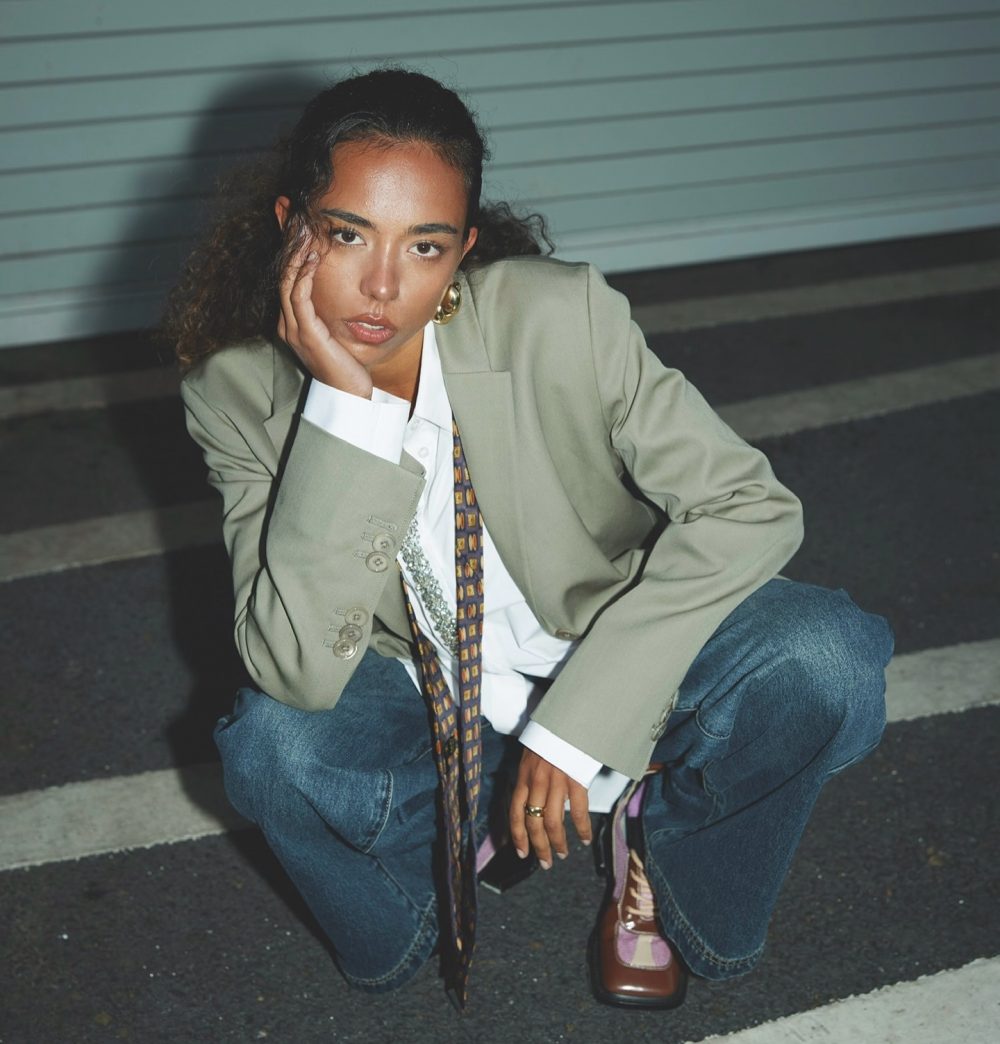 Woman crouches on a stairway at night, wearing a beige blazer, white shirt, patterned tie, and jeans, with her hand resting on her cheek and gold hoop earrings.