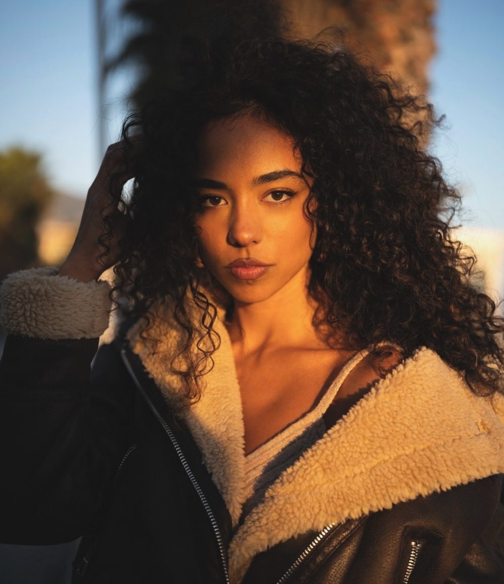 Portrait of a young woman with dark, curly hair wearing a tan shearling jacket, looking directly at the camera in golden hour light.
