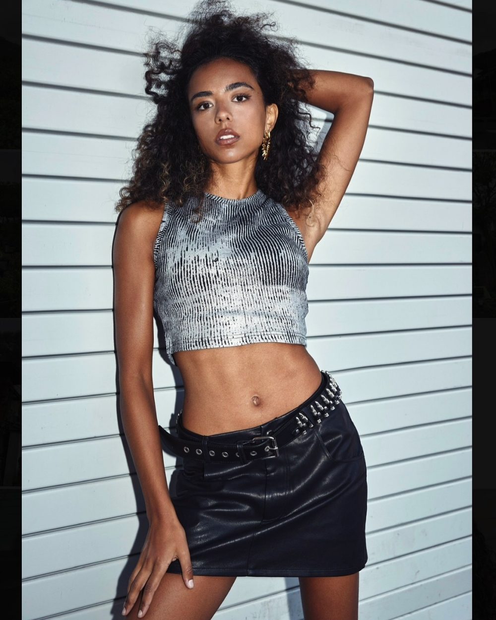 Model with curly hair wearing a metallic silver crop top and black studded belt skirt, posing against white horizontal siding.