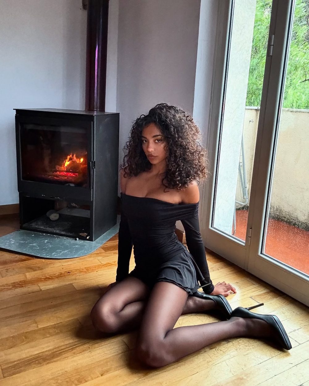 Woman kneeling on a wooden floor beside a lit fireplace, wearing an off-shoulder black dress and sheer black tights with high heels.