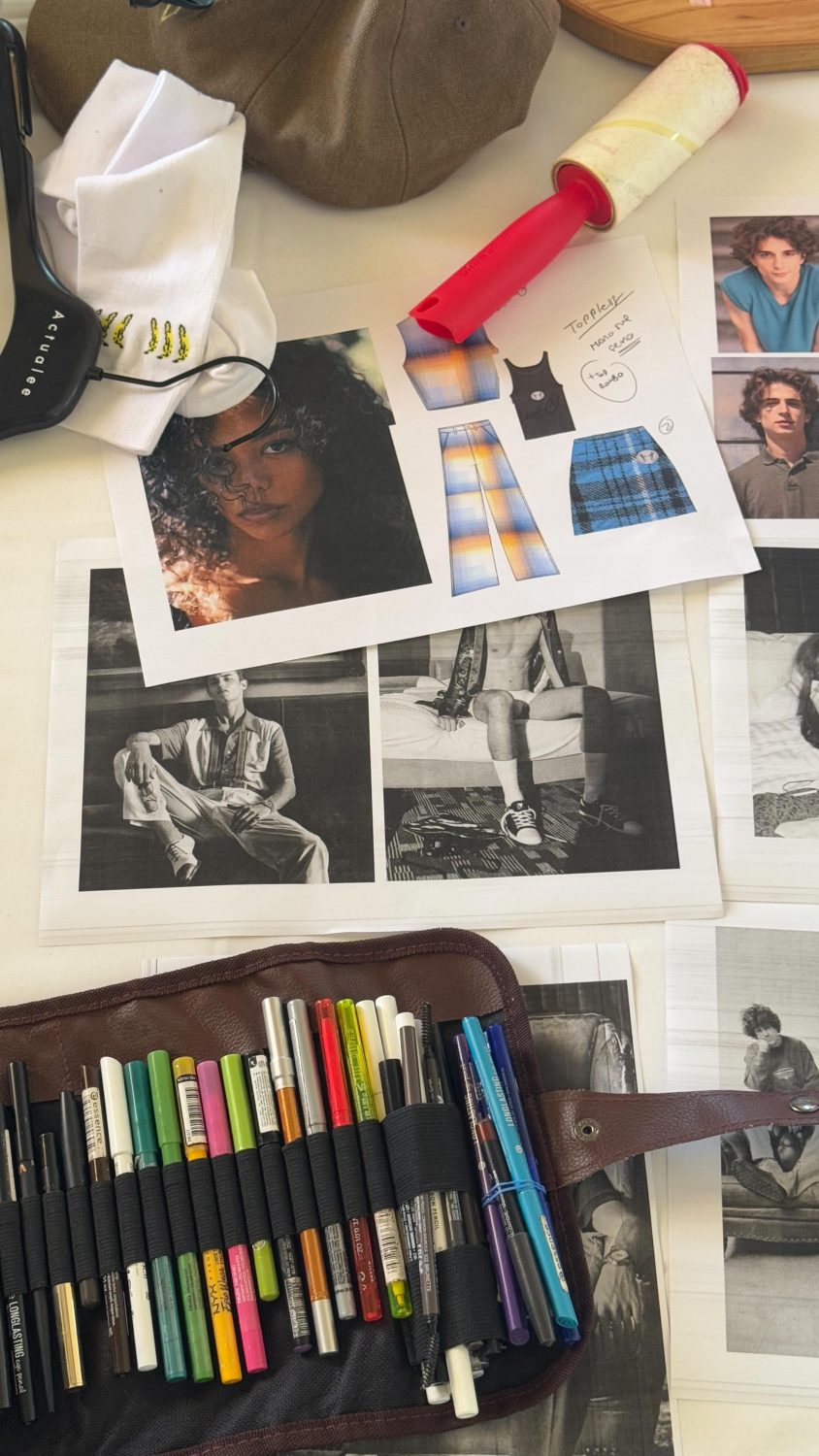 Designer’s workspace with a brown bag, sketches, fashion photos, and a case of colorful markers on a desk.