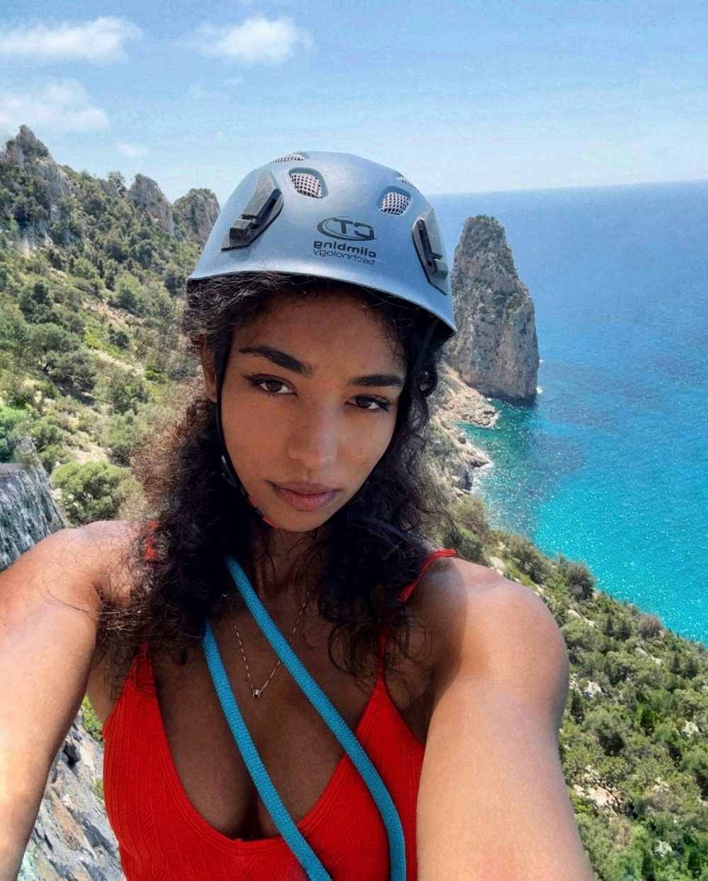 Selfie of a woman wearing a light blue climbing helmet on a rocky coastal cliff with turquoise sea in the background.
