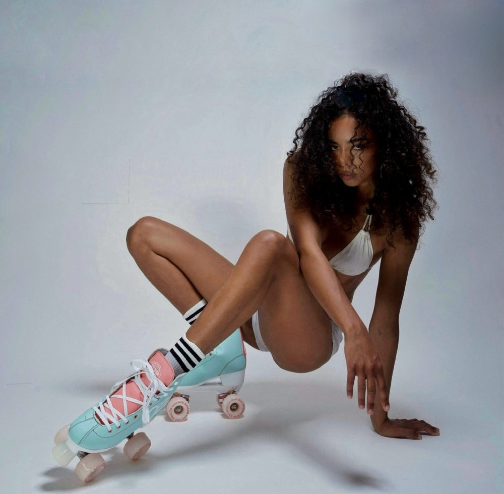 Woman with curly hair in a white bikini top crouching on a white studio floor wearing pastel roller skates with pink and teal accents.