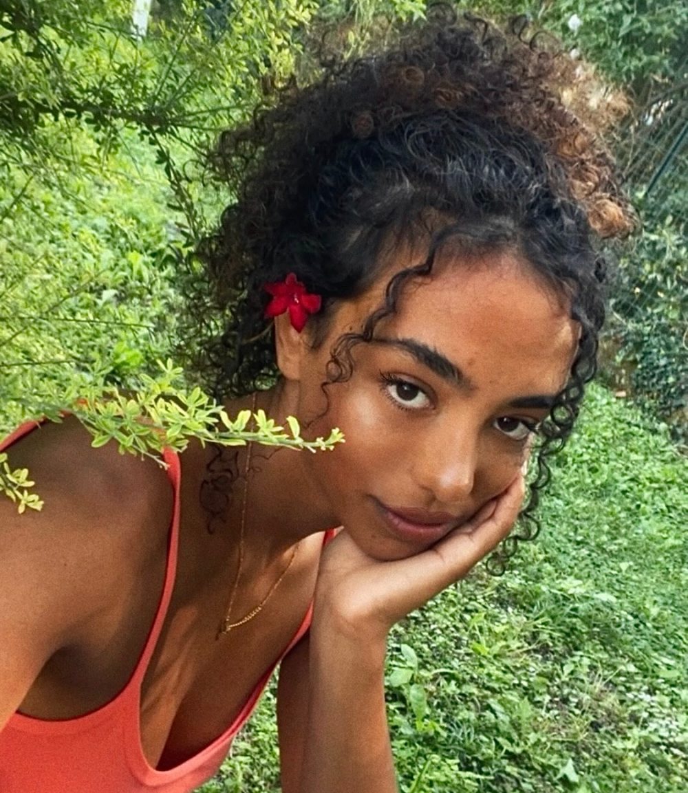 Young woman with curly hair wearing a red/orange tank top, resting her hand on her chin outdoors among greenery, a red flower in her hair.