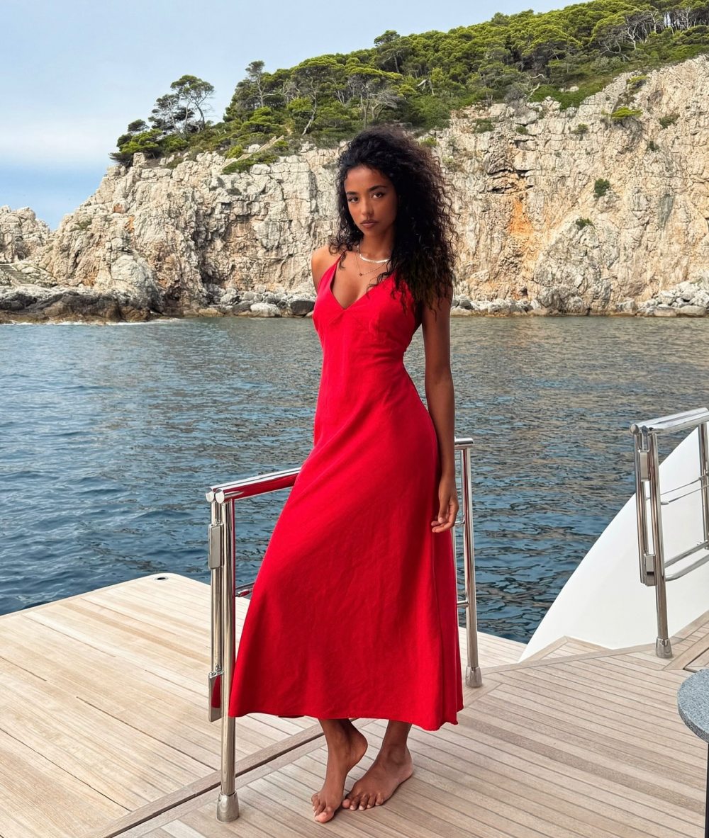 Woman in a bright red dress standing barefoot on a boat deck by rocky shoreline and blue water.