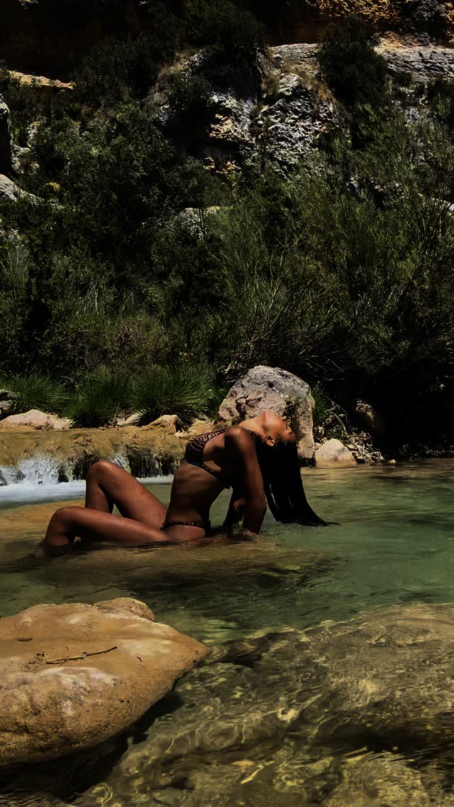 Woman in a black bikini reclining in a shallow rocky pool with green vegetation behind her.