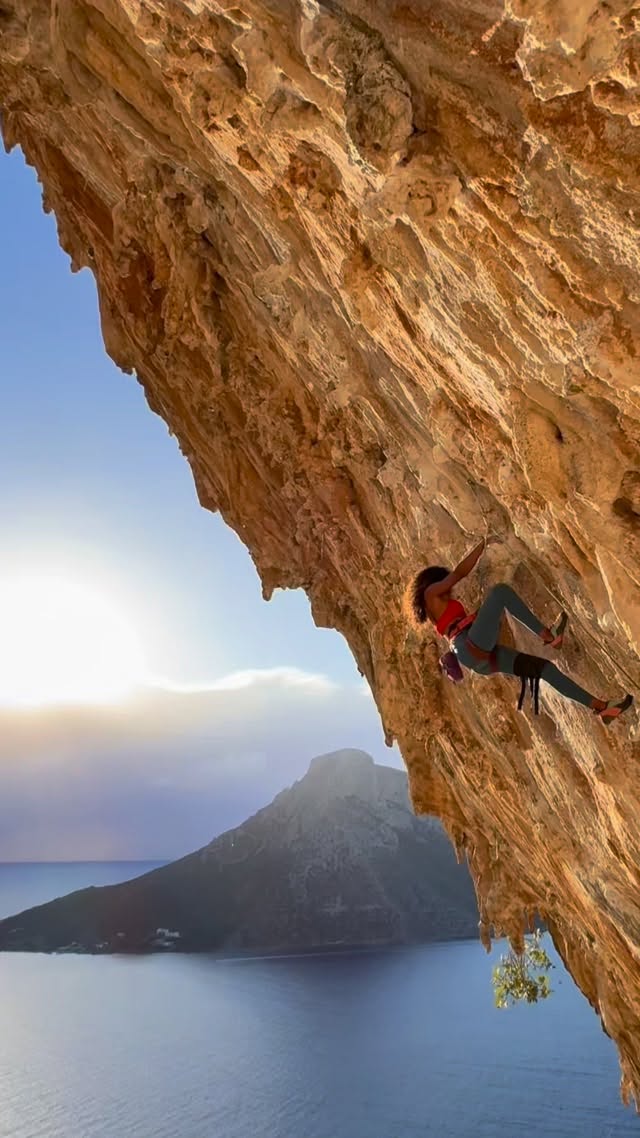 Climber suspended on a steep rocky cliff above the sea at sunset, with rugged island in the distance.