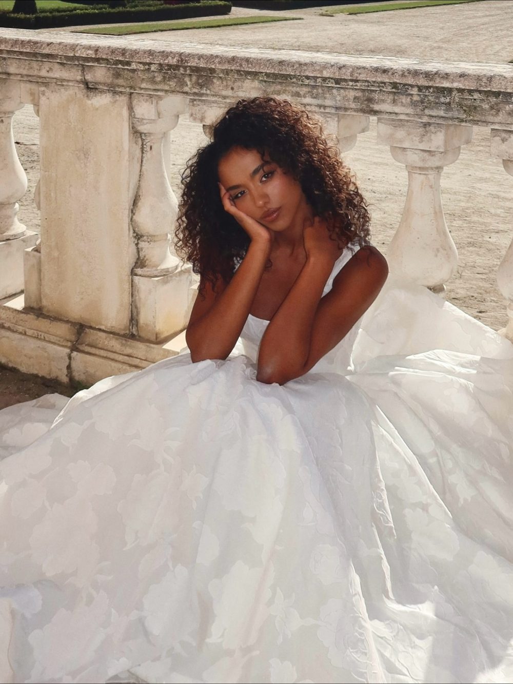 Woman in a white wedding dress sits beside a stone balustrade, resting her head on her hand.