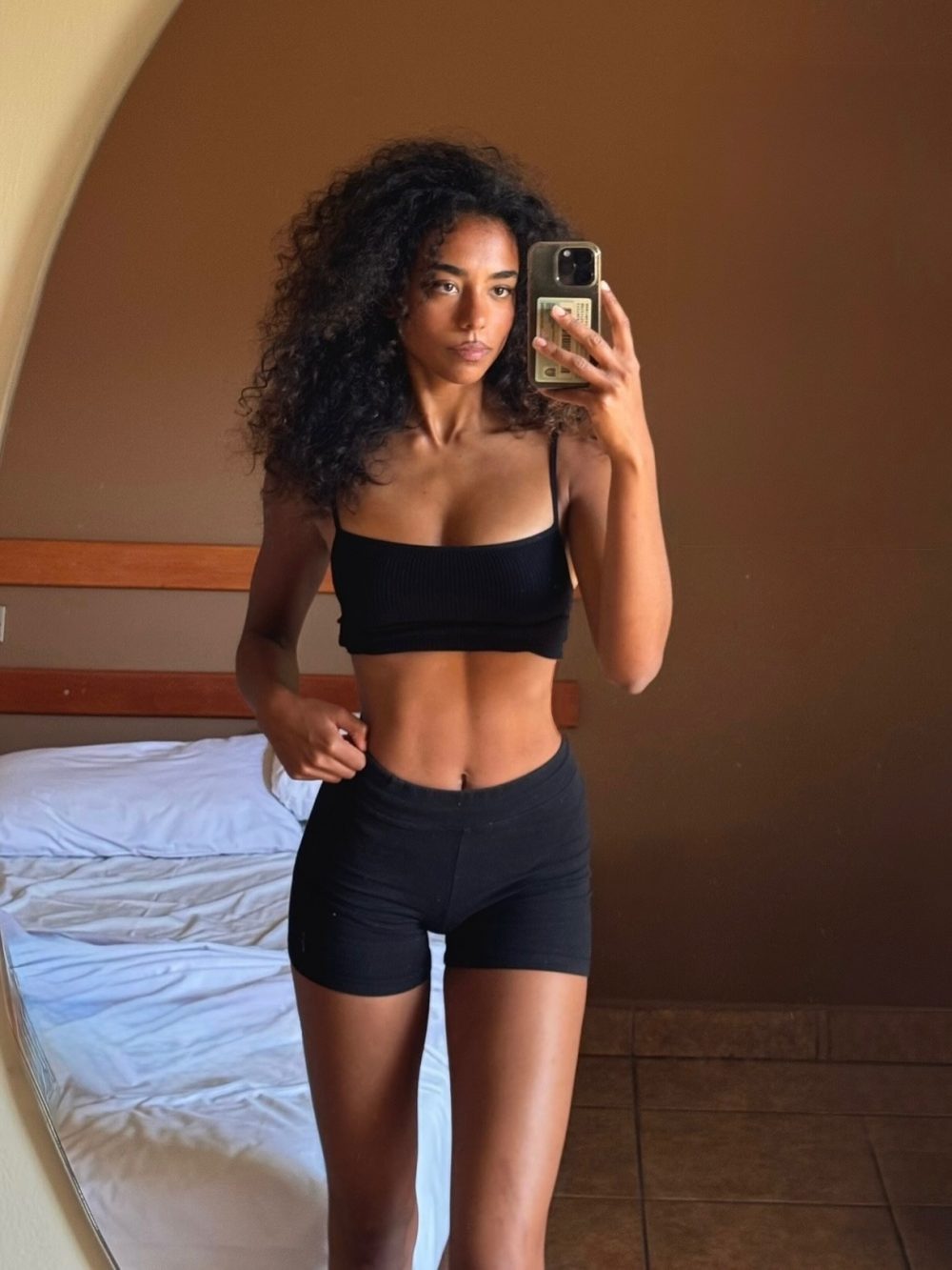 Woman in a black sports bra and black shorts taking a mirror selfie in a bedroom or hotel room, holding her phone up to capture her reflection.