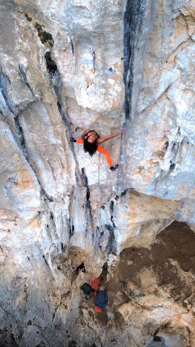 Climber in an orange outfit hangs from a vertical rock face, ropeed in, with a belayer at the base managing the rope below.