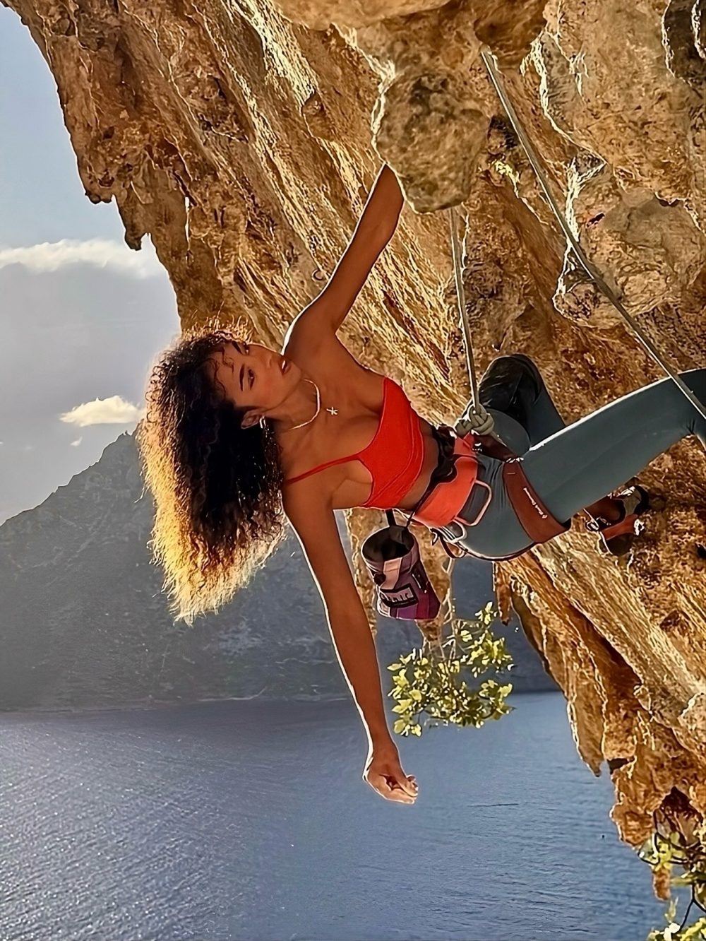 Climber in a red top ascends a rocky cliff, secured with a rope and harness above a blue body of water.