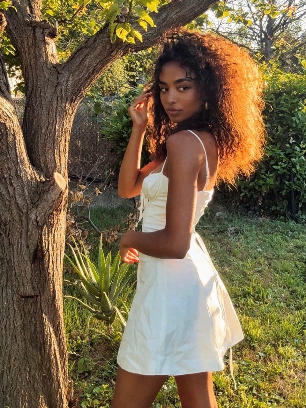 Young woman in a white sundress standing by a tree in a sunlit garden, looking at the camera.