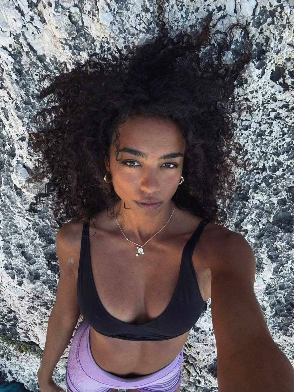 Woman with curly hair wearing a black bikini top, standing against a rough rocky backdrop, looking at the camera.