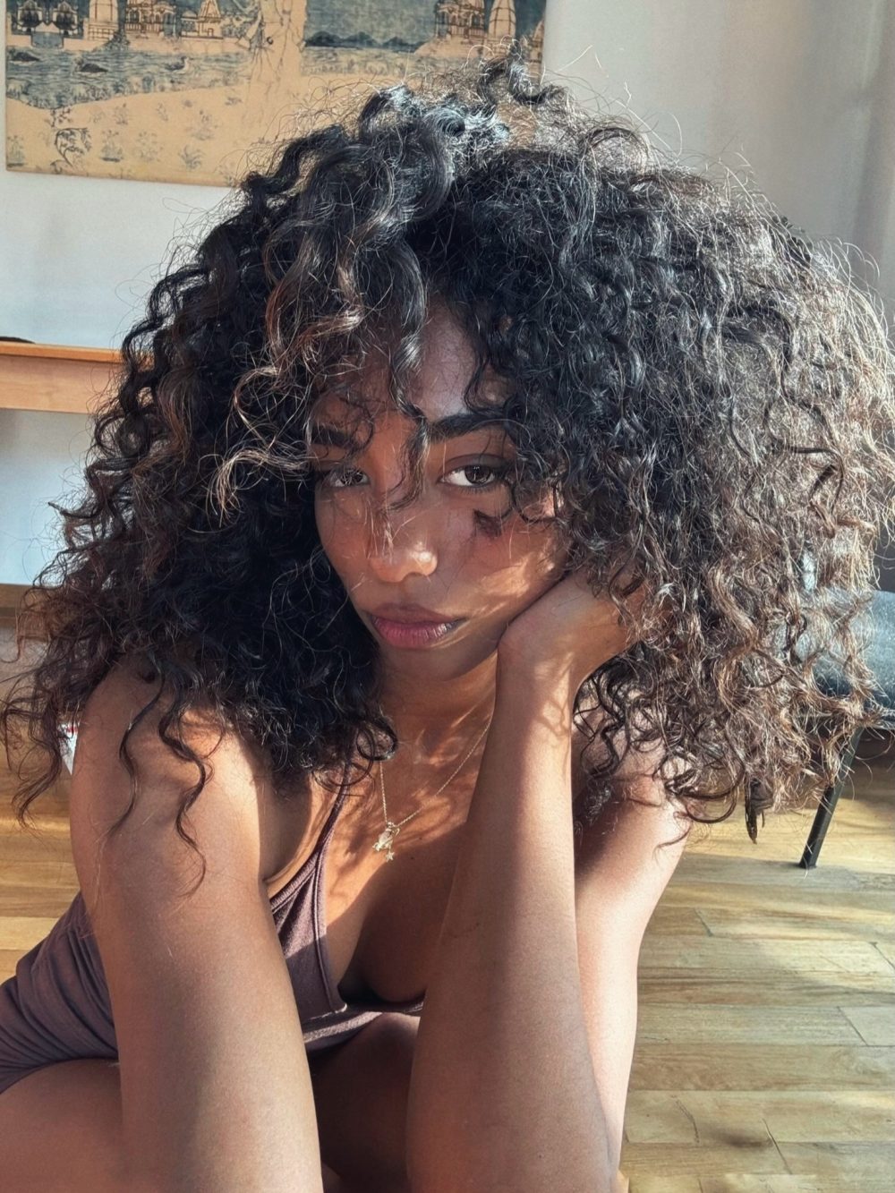 Young woman with long curly hair resting her chin on her hands, looking at the camera indoors near a wooden floor and wall art in the background.
