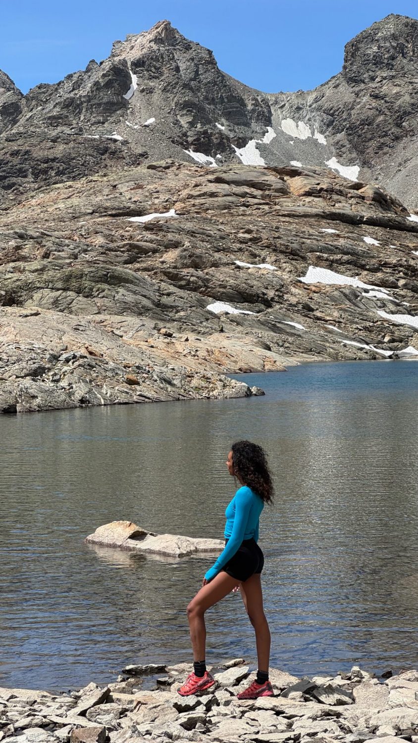 Woman in a bright blue long-sleeve shirt and black shorts stands on rocky shore beside a calm lake, with rugged mountain peaks behind.
