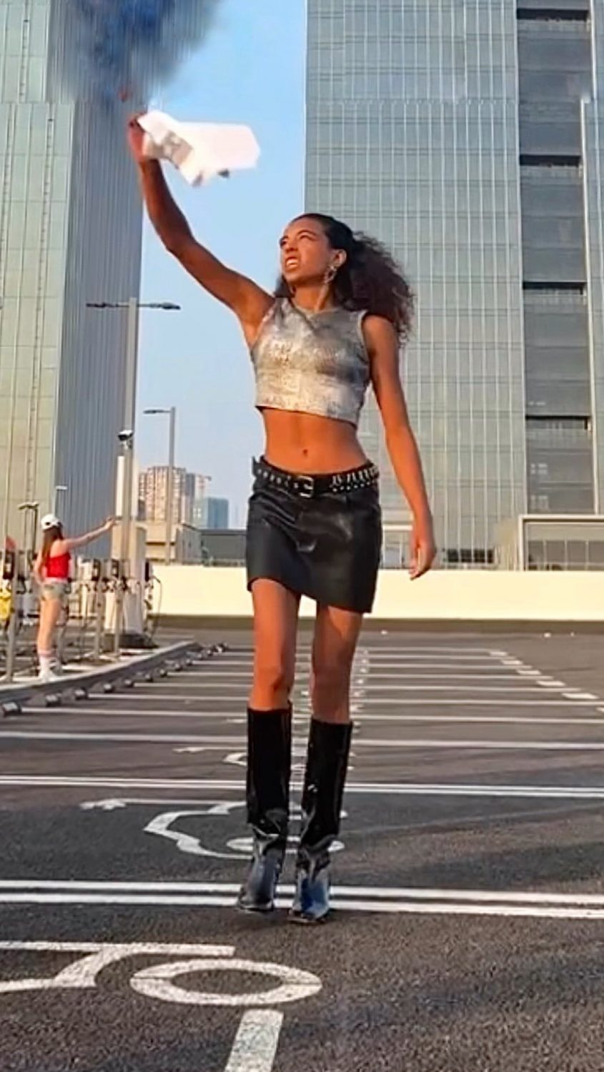 Woman in a metallic silver crop top and black leather mini skirt raises a white cloth in a rooftop parking lot with tall glass buildings behind her.