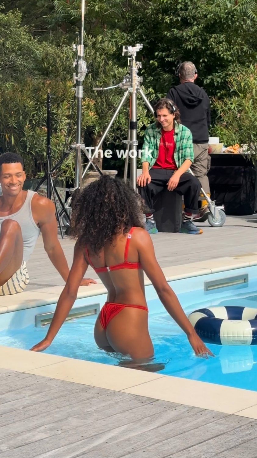 Behind-the-scenes pool shoot: a woman in a red bikini steps into a pool while two people and camera gear are visible in the background; the image includes the caption 'The work'.