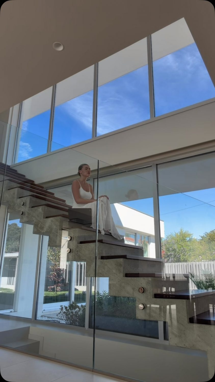 Woman in white dress seated on a glass-and-concrete staircase inside a bright modern home with large windows and blue sky outside.