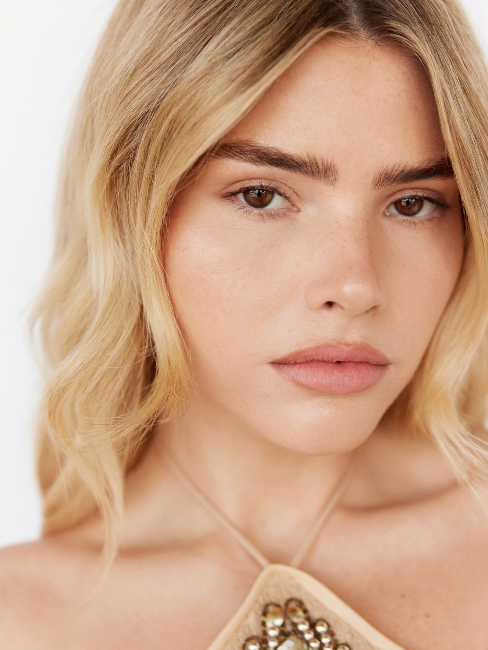 Close-up portrait of a woman with blonde hair, brown eyes, and pink lips, showing a beaded neckline.
