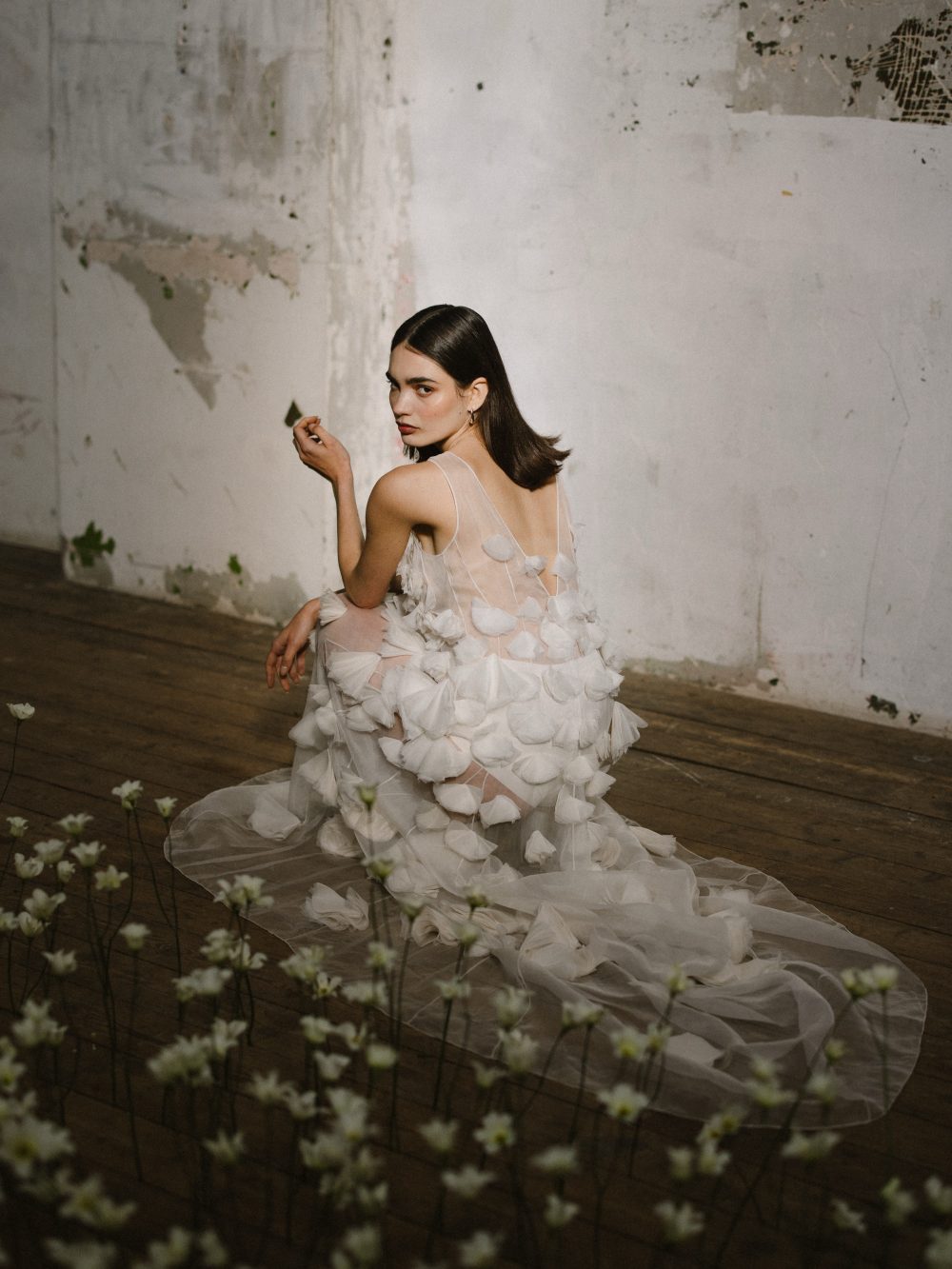 Woman in a sheer white petal-themed gown sitting on a wooden floor in a worn, white-washed room; foreground flowers.