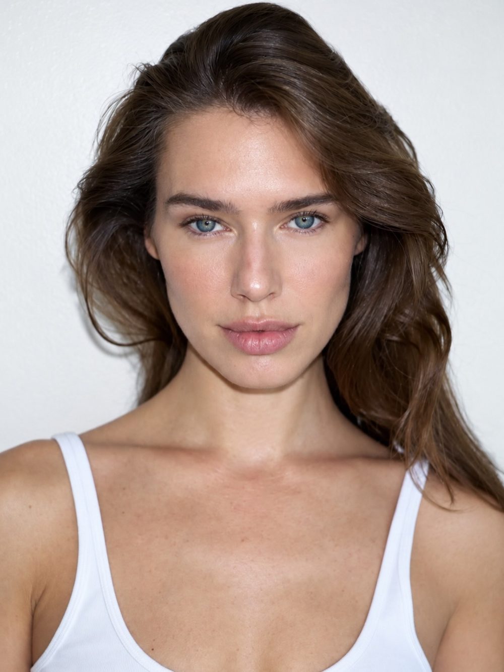Close-up portrait of a woman with blue eyes and brown hair wearing a white tank top against a light background.