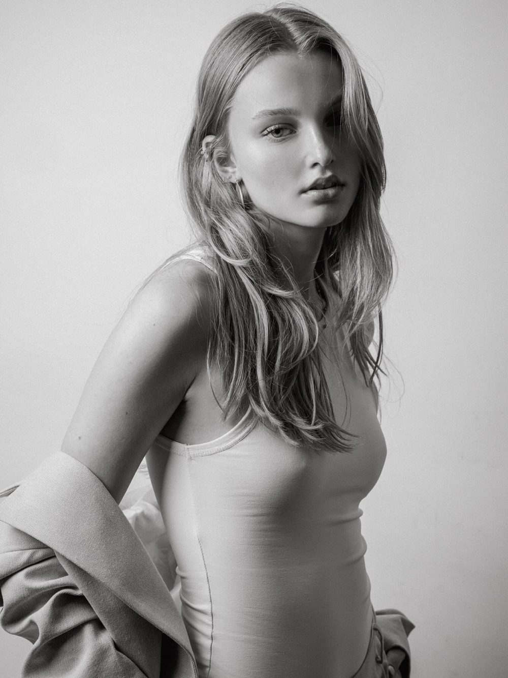 Black-and-white portrait of a young woman with long, wavy hair wearing a tank top, with a jacket draped over her arm, looking at the camera.
