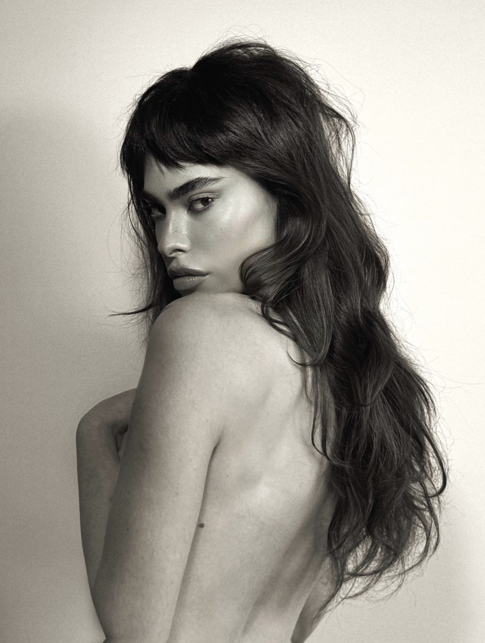 Monochrome portrait of a topless woman with long dark hair, facing left and looking over her shoulder.
