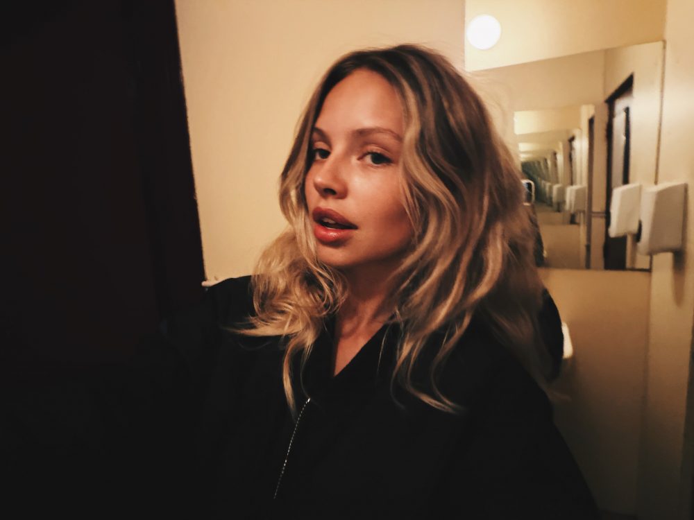 Close-up selfie of a woman with wavy blonde hair wearing black, standing in a hallway with mirrors in the background.